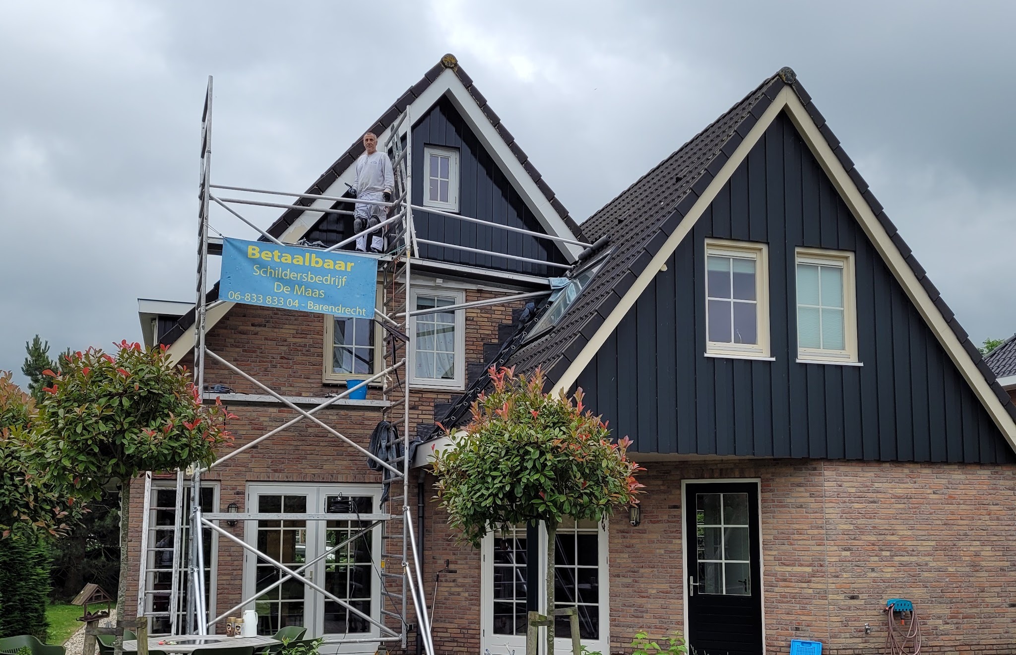 Rolsteiger met het blauwe banner Betaalbaar Schildersbedrijf De Maas aan een woning met twee puntgevels, een schilder bovenop