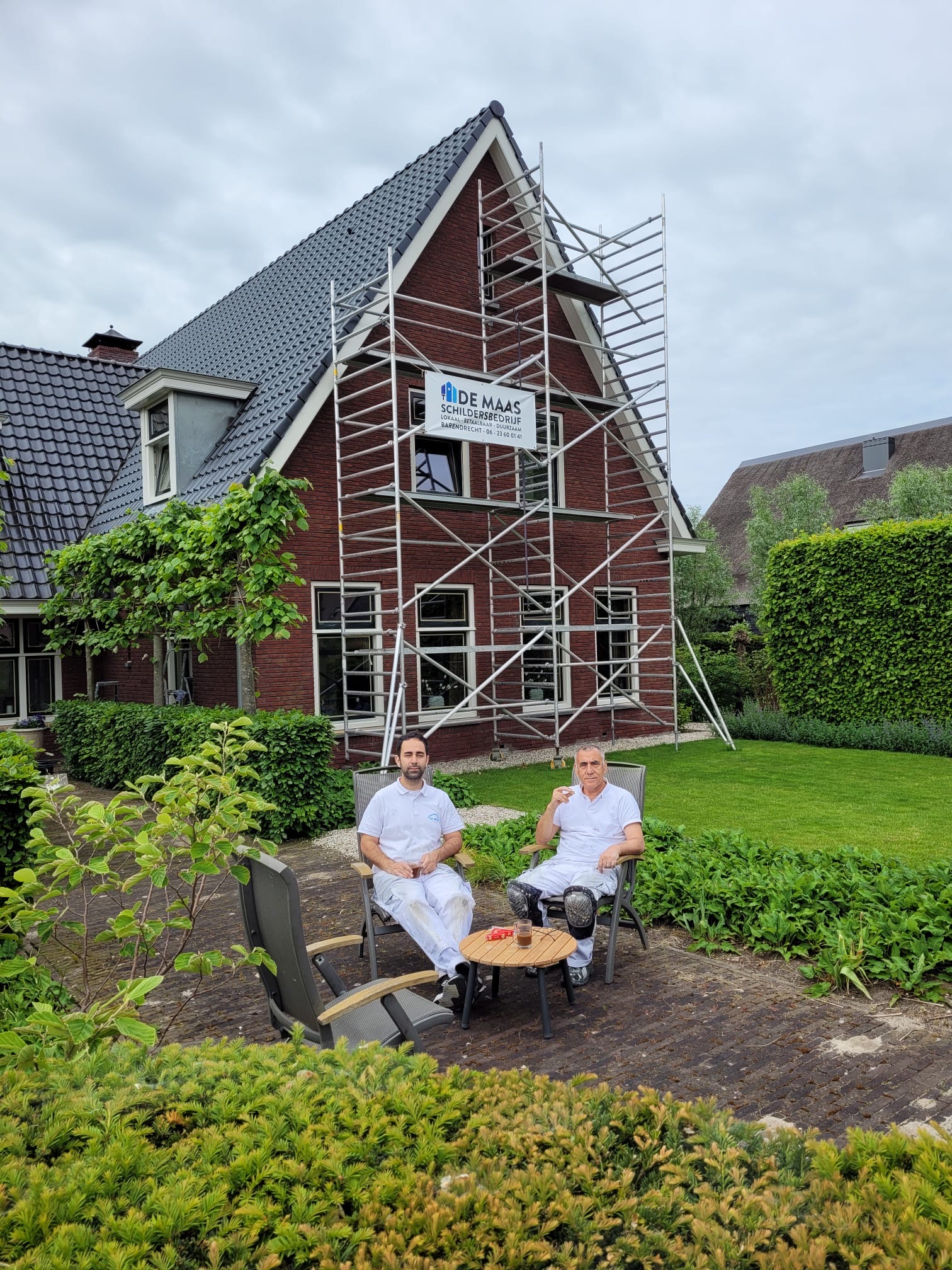 Hashem en Payam Belanj in witte werkkleding op tuinstoelen voor een woning met hun steiger en banner